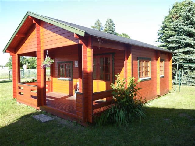 Ein kleines, rotes Gartenhaus aus Holz mit Veranda, umgeben von einem gepflegten Rasen und verschiedenen Pflanzen im Grunewald.