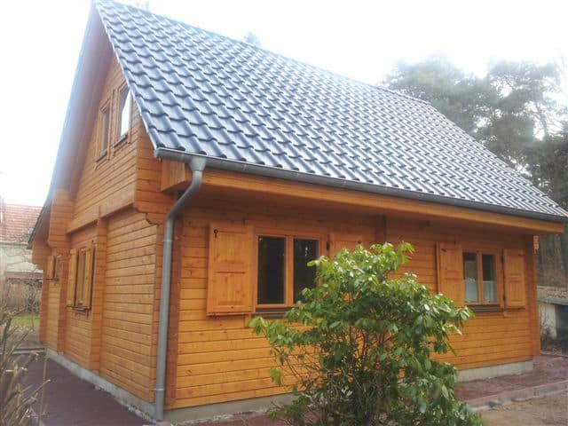 Ein hochwertiges Holzhaus mit steilem Ziegeldach und Fensterläden, umgeben von Vegetation und einem klaren Himmel darüber.