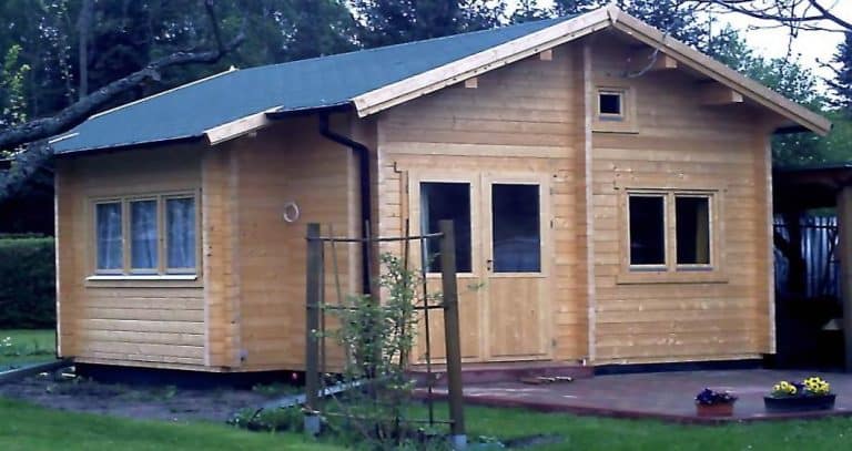 Eine kleine Holzhütte mit schrägem Dach und überdachter Veranda, umgeben von einem Rasenhof mit einem jungen Baum, der von Pfählen gestützt wird. Dieses Gartenhaus erinnert an die in M