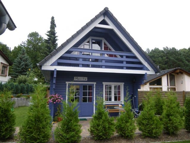 Ein blaues, zweistöckiges Ferienhaus Rosenhag mit weißen Verzierungen und A-Rahmen-Design, umgeben von viel Grün und einem gepflegten Garten.