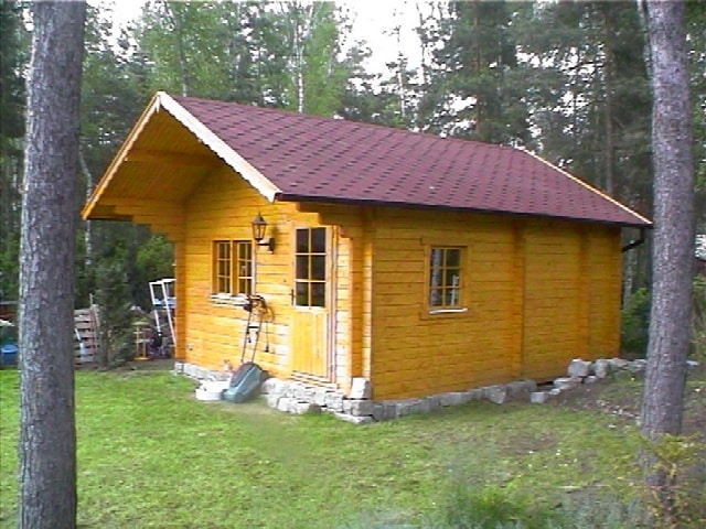 Ein kleines hölzernes Gartenhaus mit rotem Dach, umgeben von Bäumen in einer Waldgegend.